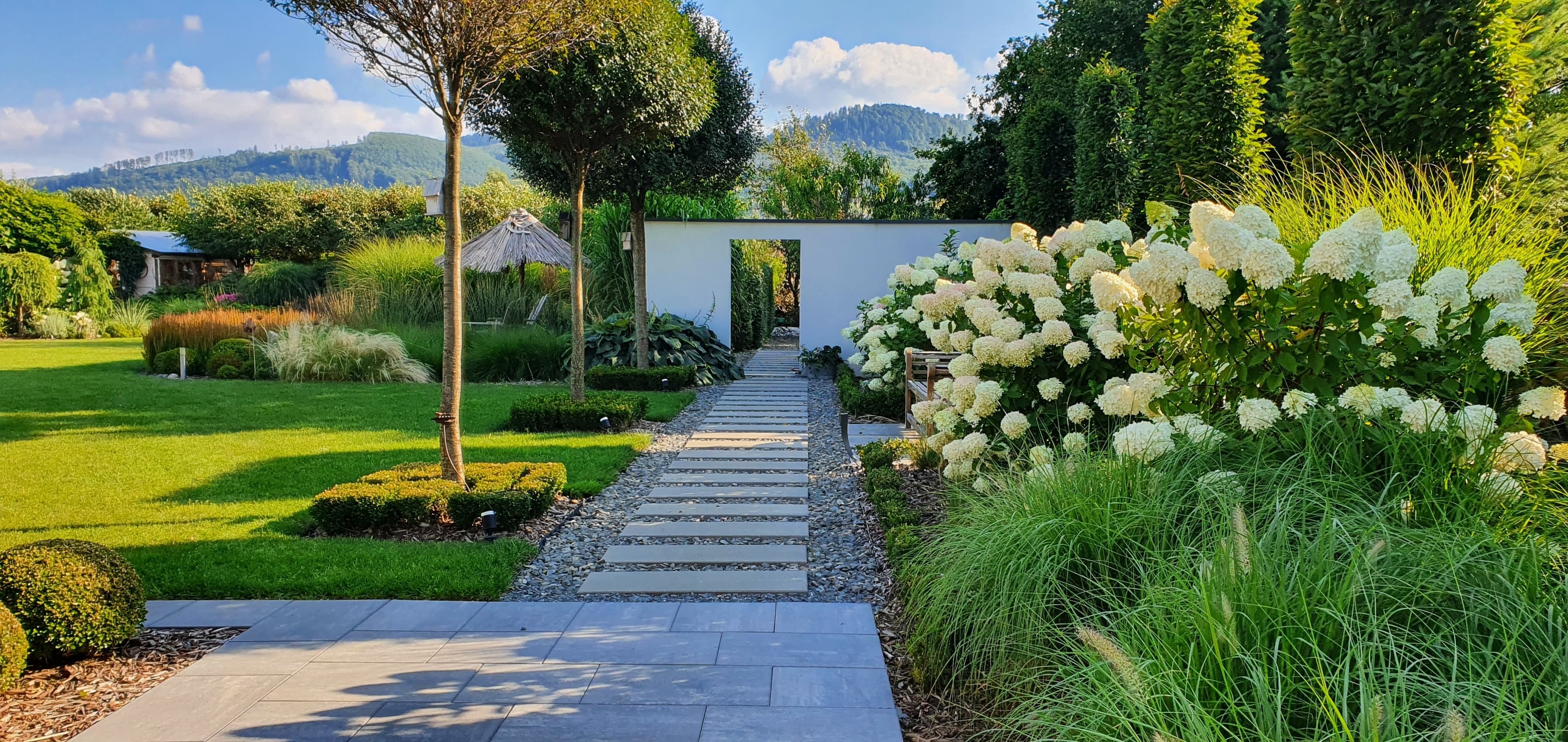 Moderner Garten mit Terrasse, Trittsteinweg und weiß blühenden Hortensien vor bergiger Landschaft.